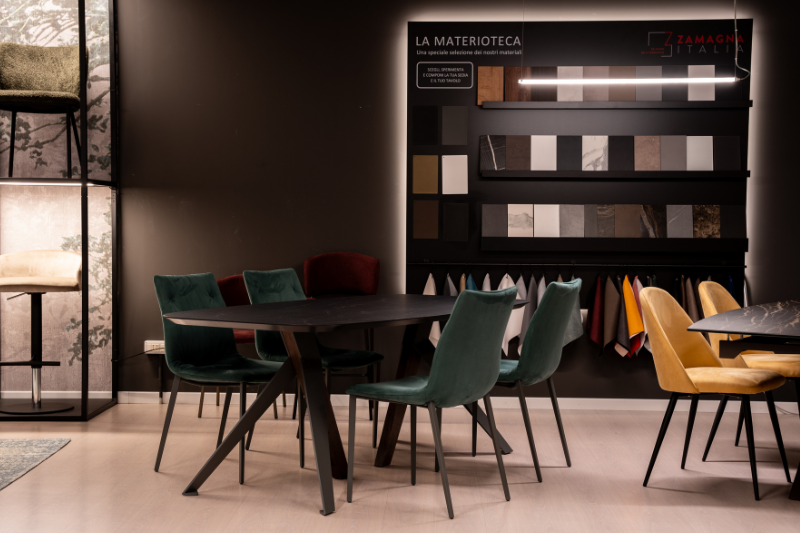 Showroom con tavolo in ceramica scura, sedie in velluto verde e giallo e parete materioteca con campioni di materiali